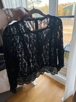 Svart spetsblus med lång ärm - Säljer en svart, transparent spetsblus med långärmad design och rund halsringning. Blusen har fina spetsdetaljer och ett dekorativt avslut nertill.