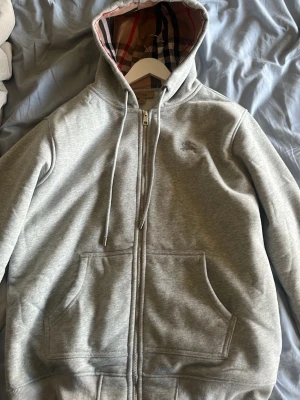 Grå zip-hoodie från Burberry - Snygg grå hoodie med dragkedja från Burberry. Klassisk huva med det ikoniska rutmönstret på insidan, dragsnören och stora fickor framtill. Perfekt för en chill och stilren look.