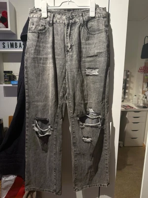 Grå slitna raka jeans - Säljer ett par grå raka jeans med slitningar och hål på båda benen. Jeansen har hög midja, klassiska fem fickor och är tillverkade i bomull. Perfekta för en avslappnad och trendig look.