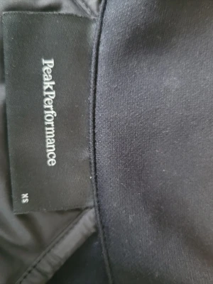 Svart dunjacka Peak Performance XS - Snygg svart dunjacka från Peak Performance i storlek XS. Jackan har quiltad panel på ryggen och släta partier på sidorna och ärmarna. Perfekt för kalla vinterdagar och har en modern, sportig look.