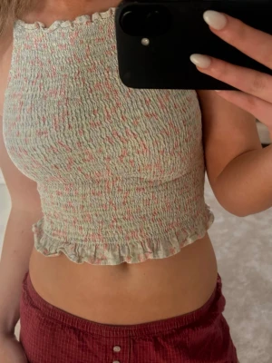Ljus croppad smockad linnetopp - Söt croppad linnetopp i ljusa pastellfärger med smockat tyg och volangkant nertill. Toppen är ärmlös och har tunna axelband som knyts i nacken. Perfekt för varma dagar och ger en somrig vibe.