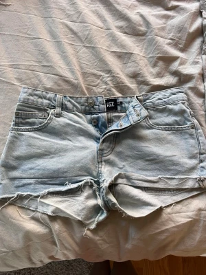 Ljusblå denimshorts från 157 - Säljer ett par ljusblå denimshorts från 157 med råa, fransiga kanter och klassisk femficksdesign. Shortsen har hög midja och knappar framtill. Perfekta för sommarens alla äventyr!