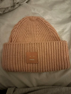 Beige stickad mössa från Acne Studios - Säljer en beige stickad mössa från Acne Studios med bred uppvikt kant och det klassiska ansiktsloggan framtill. Mössan är ribbstickad och har en mjuk, chunky känsla som håller dig varm och snygg under kalla dagar.