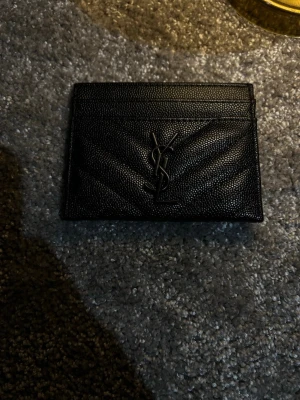 Svart YSL kortfodral i läder - Snyggt svart kortfodral från Yves Saint Laurent i präglat läder med klassisk YSL-logga i metall framtill. Fodralet har flera kortfack och stilrena sömmar i chevronmönster. Perfekt för dig som vill ha en lyxig och stilren accessoar.