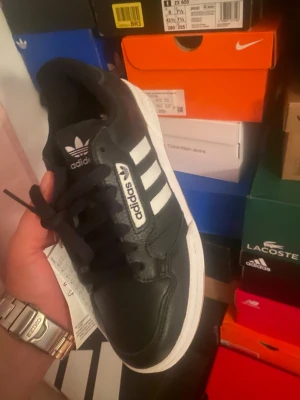 Svarta Adidas sneakers med vita ränder - Svarta Adidas sneakers med klassiska vita ränder på sidorna och vit sula. Modellen har svart ovandel i läderlook, svart plös med Adidas-logga och svarta skosnören. Perfekt för dig som gillar en clean och sportig stil.