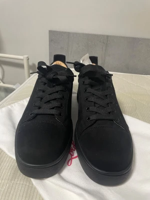 Svarta Louboutin sneakers med röd sula - Snygga svarta sneakers från Christian Louboutin med klassisk röd sula och diskret logga undertill. Skorna har en stilren design i mocka och textil, svarta skosnören och låg profil. Perfekta för dig som vill sticka ut med exklusiva detaljer. Utan kartong endast påse