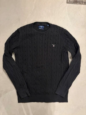 Svart kabelstickad tröja från Gant - Kabelstickad svart tröja från Gant med rund halsringning och diskret broderad logotyp på bröstet. Tröjan har långa ärmar och ribbade muddar vid ärmslut och nederkant. Perfekt för en stilren och klassisk look.