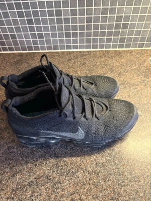 Svarta Nike Air Vapormax sneakers - Snygga helsvarta Nike Air Vapormax sneakers med stickad ovandel i mesh och genomskinlig sula. Skorna har klassisk Nike-logga på sidan, rundad tå och elastiska skosnören. Perfekta för dig som gillar sportig och modern stil.