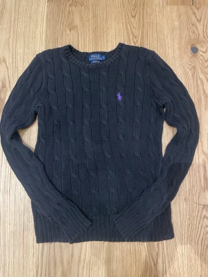 Mörkgrå kabelstickad tröja Polo Ralph Lauren storlek S - Kabelstickad mörkgrå tröja från Polo Ralph Lauren med lila broderad logga på bröstet. Tröjan har rund halsringning, långa ärmar och ribbade muddar. Jag säljer pga jag inte använder denna tröjan längre. Skriv vid minsta funderingar. MVH Tim.