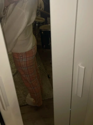 Rutiga pyjamasbyxor i orange och rosa - Säljer ett par mysiga pyjamasbyxor med rutigt mönster i orange, rosa och vitt. Byxorna har resår i midjan och är perfekta för chill hemma. Materialet ser ut att vara mjukt och bekvämt, troligen bomull eller bomullsblandning.