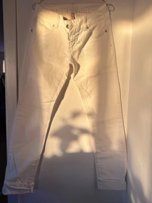 Vita raka jeans med hög midja - Säljer ett par vita jeans med rak passform och hög midja. Klassisk femficksmodell med bälteshällor och knappgylf. Jeansen är i ett tjockare bomullstyg och har enkel, stilren design utan slitningar eller dekorationer.