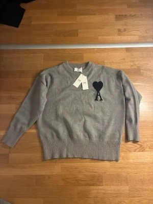 Grå stickad tröja från Ami Paris - M-L. Snygg grå stickad tröja från Ami Paris med svart hjärta och bokstaven A broderat på bröstet. Tröjan har ribbade muddar vid ärmslut och nederkant samt rund halsringning. Perfekt för dig som gillar stilrena och trendiga plagg.