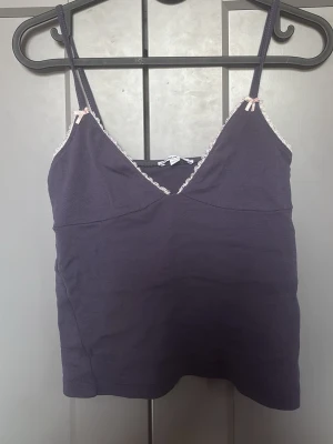 Lila  linnetopp från Subdued - Supersöt Lila linnetopp från Subdued med smala axelband, v-ringning och spetsdetaljer längs kanten. Små ljusrosa rosetter vid axelbanden ger en gullig touch. Perfekt att styla med jeans eller kjol.