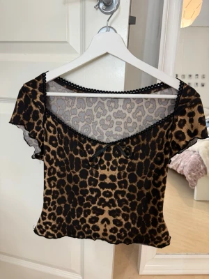 Leopardmönstrad croppad topp - Snygg croppad topp med leopardmönster och korta ärmar. Toppen har en liten rosett framtill och dekorativ spetskant vid urringningen. 🖤