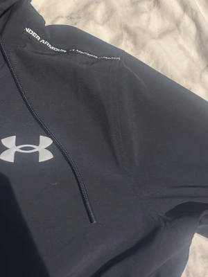 Svart hoodie från Under Armour - Snygg svart hoodie från Under Armour med stor logga på bröstet och justerbar dragsko i huvan. Tillverkad i ett mjukt och funktionellt material som passar perfekt till träning eller chill. Klassisk passform och diskret branding på huvan.