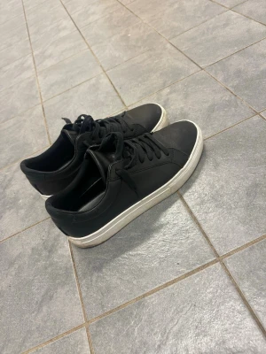 Svarta Vagabonds skor - Stilrena svarta sneakers från Cropp med klassisk låg profil och vit sula. Skorna har svarta skosnören och ovandel i syntetmaterial som ger en clean look. Perfekta för dig som gillar minimalistisk streetstyle.