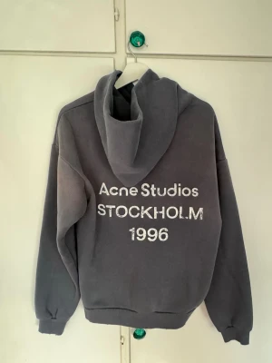 Grå hoodie Acne Studios Stockholm - Säljer en grå hoodie från Acne Studios med stor vit text på ryggen: 'Acne Studios STOCKHOLM 1996'. Hoodien har huva och ribbade muddar vid ärmslut och nederkant. Perfekt för dig som gillar streetwear och vill ha en stilren look.