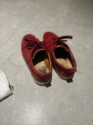 Röda sneakers med nitar från Louboutin kan möjligen prutas - Unika röda sneakers i mocka från Christian Louboutin med coola nitar på tån och klassiska röda sulor. Skorna har snörning och låg profil som ger en snygg streetwear-vibe. Perfekta för dig som vill sticka ut med exklusiva detaljer och lyxig känsla.