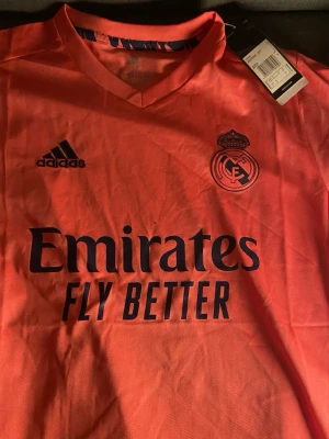 Real Madrid orange matchtröja Adidas - Snygg orange matchtröja från Adidas för Real Madrid med klubbmärke och sponsortryck på bröstet. Tröjan har V-ringning och korta ärmar, perfekt för fotbollsträning eller match. Materialet är lätt och andas bra.