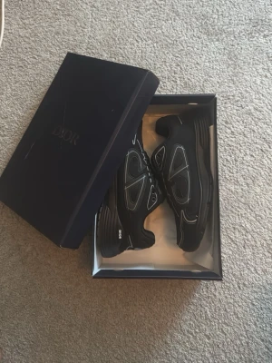 Svarta Dior sneakers med silverdetaljer - Säljer ett par svarta Dior sneakers med coola silverdetaljer och mesh-paneler. Skorna har en chunky sula och klassisk låg profil. Märkeslogga på tungan och sidan. Perfekt för dig som vill ha en exklusiv och trendig look.