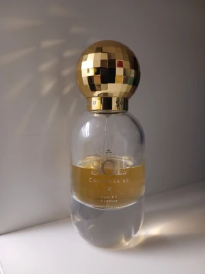 Sol de Janeiro Cheirosa '62 EdP - Sol de Janeiro Cheirosa '62 Eau de Parfum med en unik rund flaska och en glansig, guldmönstrad kork. Flaskan är genomskinlig med guldfärgad etikett och parfymen har en varm, gyllene ton. Perfekt för dig som gillar lyxig och iögonfallande design.