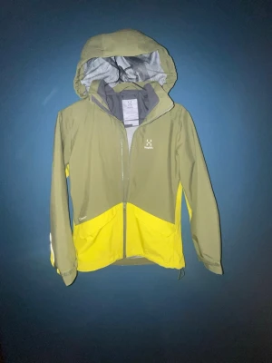 Grön och och ljusgrön regnjacka från Haglöfs - Snygg regnjacka från Haglöfs i olivgrönt och gult med hel dragkedja och avtagbar huva. Jackan har meshfoder på insidan och logga på bröstet. Perfekt för regniga dagar och outdoor-äventyr. Tillverkad i slitstarkt, vattenavvisande material. Nästan helt onanänd.