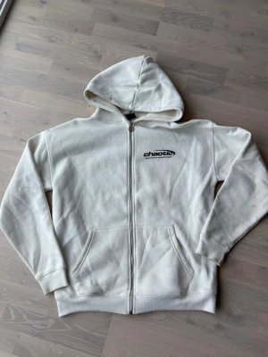 Vit zip hoodie från Chaotic - Vit hoodie med dragkedja från Chaotic. Tryck med logga på bröstet och stort svart tryck på ryggen med texten 'Aesthetic Awareness From Outta Space'. Mjuk bomullskänsla, två fickor fram och klassisk huva. Perfekt för en avslappnad streetwear-look.