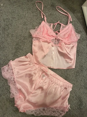 Rosa satinset med spetsdetaljer - Sött tvådelat set i ljusrosa satin med spetsdetaljer. Toppen har tunna axelband, spets vid bysten och dekorativa band framtill. Shortsen har elastisk midja, spetskant och liten rosett. Perfekt för en mysig kväll hemma.