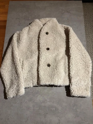 Krämvit teddyjacka med knappar - HELT OANVÄND MED PRISLAPP KVAR! Mysig teddyjacka ifrån gina tricot i krämvit färg med tre stora knappar framtill. Jackan har en boxig passform och är tillverkad i fluffigt teddy-material som ger en trendig look. Perfekt för att hålla värmen under kyliga dagar. 