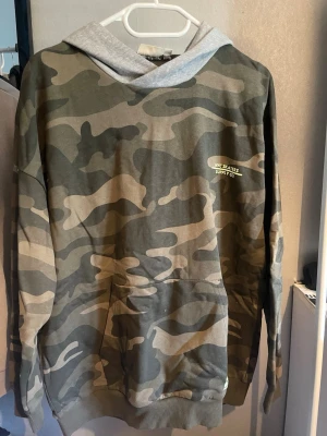 Camo hoodie från NYC Skaters Supply Co. - Snygg hoodie med camouflagemönster i gröna och beige toner från NYC Skaters Supply Co. Grå huva och stor magficka framtill. Perfekt för en streetwear-look och chill vardagsstil.