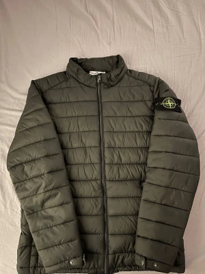 Mörkgrön pufferjacka från Stone Island - Snygg mörkgrön pufferjacka från Stone Island med klassisk patch på vänster ärm. Jackan har ståkrage, två dragkedjeförsedda fickor och quiltad design. Perfekt för kalla dagar och ger en stilren look. Materialet är lätt och isolerande. Tyvärr lite sönder där storlek står (se bild) kan fixas vid intresse.
