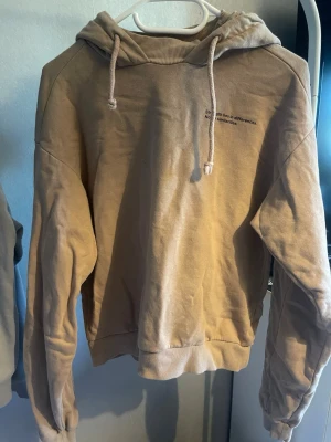 Beige hoodie med citattryck - Beige hoodie med huva och dragsko. På bröstet finns ett litet svart tryck med texten 'Strength lies in differences. Not in similarities.' Mjuk bomullskänsla och ribbade muddar vid ärmslut och nederkant. Perfekt för en avslappnad stil.