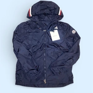 Mörkblå vindjacka från Moncler - Snygg mörkblå vindjacka från Moncler med huva och dragkedja framtill. Jackan har vita och röda detaljer på huvan samt Moncler-logga på ärmen. Tillverkad i lätt, glansigt material som skyddar mot vind. Perfekt för en sportig och trendig look.
