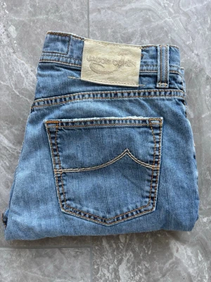 Ljusblå jeans från Jacob Cohen - Säljer ett par ljusblå jeans från Jacob Cohen med klassisk femficksmodell och raka ben. Jeansen har kontrastsömmar och en beige patch med logga bak i midjan. Tillverkade i mjukt denimtyg som ger en avslappnad look.