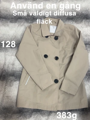 Beige trenchcoat skaljacka med svarta knappar - Stilren beige skaljacka som ser ut som en trenchcoat från Polarn O. Pyret med dubbelknäppning och svarta knappar. Klassisk krage och snedställda fickor. Perfekt för dig som gillar tidlös och enkel stil.🌸 Använd en gång. Små och väldigt diffusa fläckar. Ej tvättad. Storlek 128