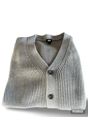 Grå ribbstickad kofta från Uniqlo - Snygg grå ribbstickad kofta från Uniqlo med v-ringning och svarta knappar framtill. Koftan har breda muddar och är gjord i ett mjukt, tjockt material som känns riktigt skönt. Perfekt att slänga över en t-shirt eller skjorta för en clean look.