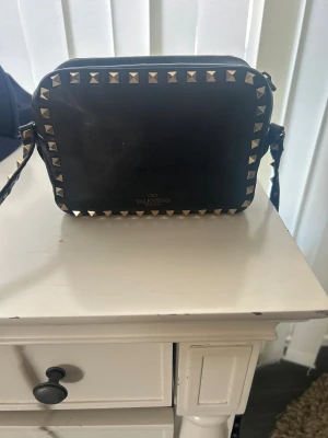 Svart axelväska Valentino Rockstud - Cool svart axelväska från Valentino med ikoniska Rockstud-nitar i guld runt kanterna och på axelremmen. Väskan är rektangulär i formen och tillverkad i slätt läder. Perfekt för dig som gillar edgy designerstil och vill sticka ut. 
