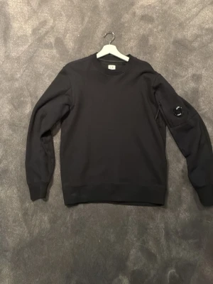 Svart sweatshirt från C.P. Company - PRISET KAN DISKUTERAS!! Svart sweatshirt med rund hals från C.P. Company. Tröjan har en ikonisk linsdetalj på ärmen och en liten ficka med dragkedja. Tillverkad i mjukt bomullsmaterial och har ribbade muddar vid ärmslut och nederkant. Hör av er vid frågor! 👍