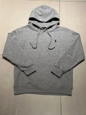 Grå hoodie från Polo Ralph Lauren - Snygg grå hoodie från Polo Ralph Lauren med klassisk huva och dragsnören. Tröjan har ribbade muddar och den ikoniska lilla svarta loggan broderad på bröstet. Perfekt för en chill och stilren look.