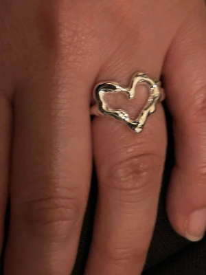 Silverfärgad hjärtformad ring - ring i silverfärgad metall med ett öppet hjärta som motiv. Designen är modern och lekfull med organiska former och en blank finish. Perfekt för dig som vill ha ett smycke med personlig touch och stil.