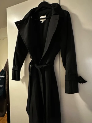 Svart trenchcoat med knytskärp - Stilren svart trenchcoat med bred krage och knytskärp i midjan. Jackan har långa ärmar med dekorativa spännen vid ärmsluten och klassisk trenchcoat-design. Perfekt för dig som vill ha en tidlös och snygg look.
