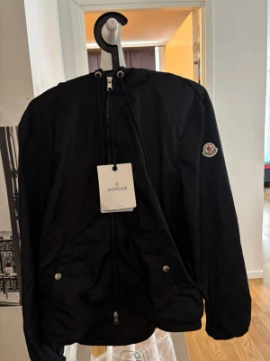 Svart vindjacka från Moncler - Snygg svart vindjacka från Moncler med huva och dragkedja framtill. Jackan har två fickor med tryckknappar och Moncler-logga på ärmen. Perfekt för blåsiga dagar och enkel att matcha med olika outfits.