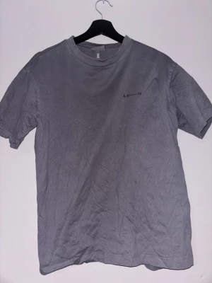 Grå t-shirt med tryck SOHO London - Grå t-shirt med svart texttryck på ryggen där det står SOHO @W1F/LDN. Liten diskret text på bröstet. Klassisk passform och rund halsringning. Perfekt för en avslappnad streetstyle-look.