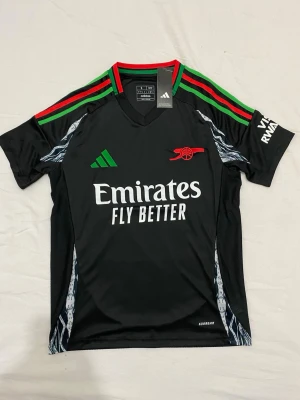 En Arsenal bortatröja 24/25 - Priset kan diskuteras. Svart Arsenal matchtröja från Adidas med gröna och röda detaljer på axlarna, vitt tryck med Emirates Fly Better på bröstet och röd Arsenal-logga. Tillverkad i lätt och ventilerande material, perfekt för fotboll eller träning.