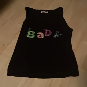Svart ärmlös topp från Louise Fd - Säljer en svart ärmlös topp från Louise Fd med färgglad text 'Baby' på framsidan. Perfekt för en avslappnad stil. Toppen är i ett mjukt material och har en bekväm passform.
