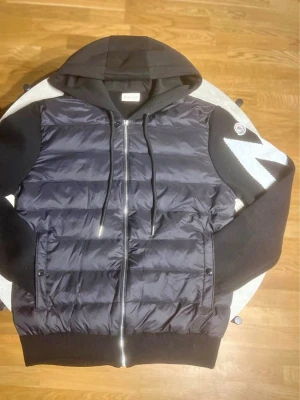 Moncler cardigan  - Svart pufferjacka från Moncler med quiltad framsida i glansigt material och stickade ärmar samt rygg. Jackan har huva med dragsko, dragkedja framtill och Moncler-logga på ena ärmen. Perfekt för kalla vinterdagar. Storlek Xl men passar L bäst så i princip storlek L