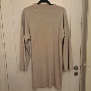 Beige långärmad klänning i bomull - Enkel och stilren beige klänning med lång ärm och rak passform. Klänningen är tillverkad i mjuk bomull och har en klassisk, avslappnad look som passar perfekt till vardags. Inga synliga detaljer eller mönster, bara ren och tidlös design.