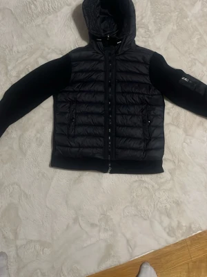 Svart pufferjacka från Moncler storlek M helt ny typ  - Snygg svart pufferjacka från Moncler med quiltad front och huva. Ärmarna är i mjukt tyg med en Moncler-logga på vänster arm och diskreta fickor med dragkedja. Perfekt för kalla dagar och ger en clean streetwear-look. Den har nfc också och det går och skanna och sånt men jag hade den i en vecka nu tänker jag sälja den jag vill köpa en annan jacka därför 