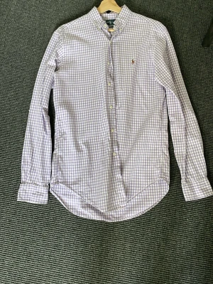 Lila rutig skjorta från Ralph Lauren - Snygg lila och vit rutig skjorta från Ralph Lauren i klassisk passform. Skjortan har knappar hela vägen, button-down krage och broderad logga på bröstet. Perfekt för dig som gillar stilrena och tidlösa plagg.