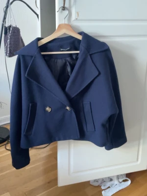 Mörkblå kort kappa med bred krage - Snygg mörkblå kappa från New collection med bred krage och dubbelknäppning framtill. Modellen är kort och har två fickor med knappar på ärmarna. Perfekt inför våren dessutom💕